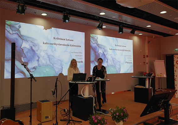 Valokuvassa tutkijat Sanna Kivjärvi ja Laura Huhtinen-Hildén seisovat lavalla,taustallaan diat, jossa lukee Kriittinen katsaus kulttuurihyvinvoinnin käsitteisiin.