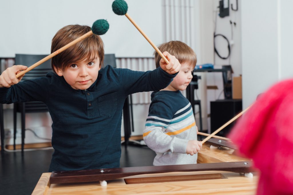 Kaksi lasta soittamassa lyömäsoittimilla. Toinen katsoo kameraan utelias hymy kasvoillaan. Two children playing percussions. One of children looks at the camera with a curious smile on his face.