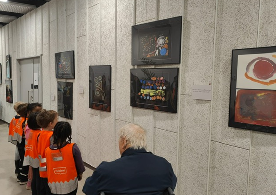 Yksi ikäihminen ja neljä lasta katsovat seinällä olevaa taidenäyttelyä. Seinällä olevat teokset ovat avaruus-aiheisia peiteväritöitä. / One elderly person and four children are looking at an art exhibition. The theme of the paintings are space.