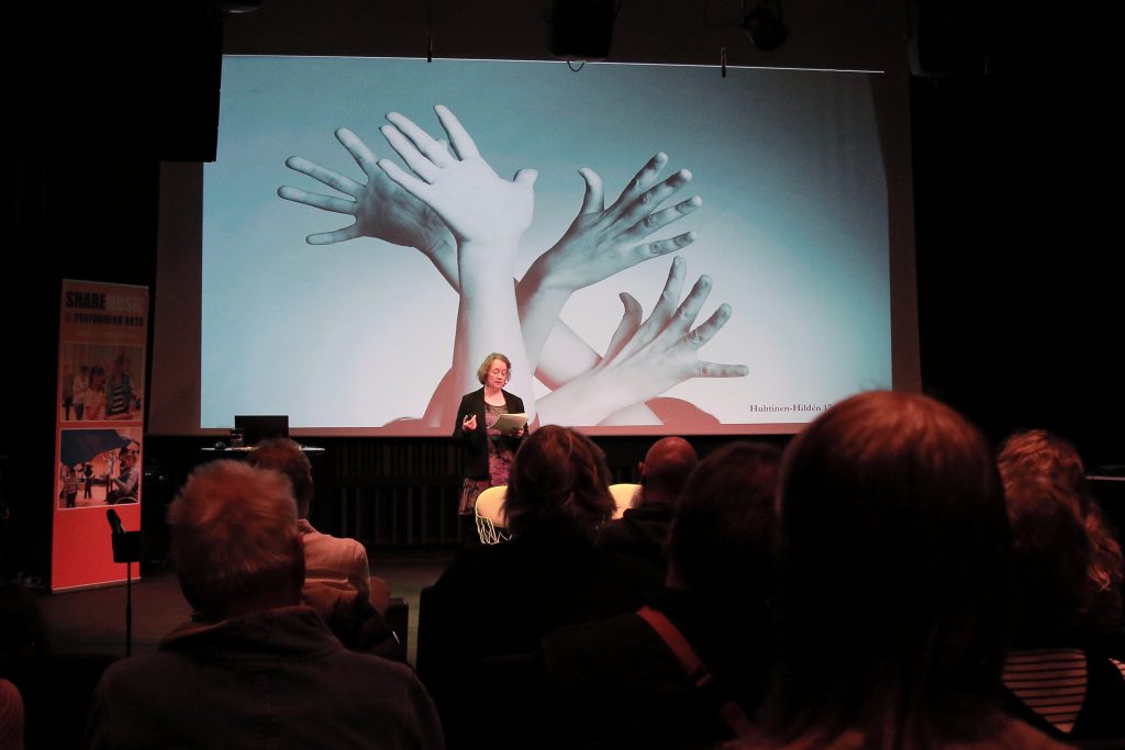 Auditorium. one person talkin in the front, behind is screen with picture of four hands.