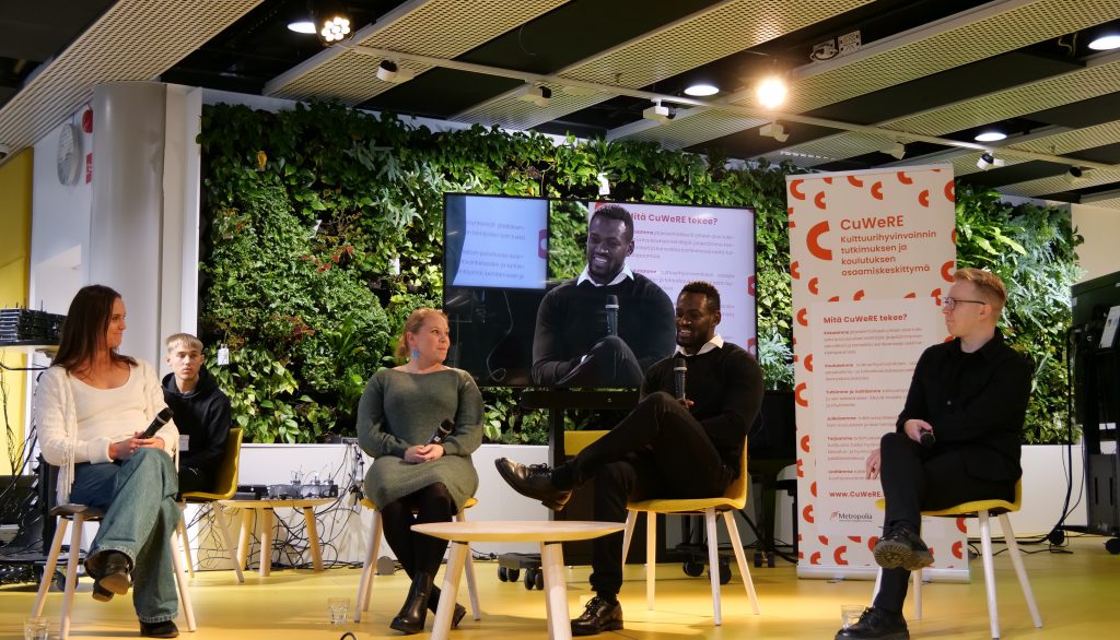 The panelists sit on chairs on the stage. In the background, CuWeRE's roll-up and video reflection of the discussion, as well as a student involved in the event production.