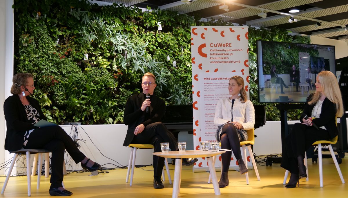 Panel discussion on a yellow stage, four panelists, one holding a microphone. In the background, a green wall and SuWeRE's roll-up and an image of the speakers projected on the video screen.