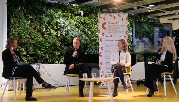 Four panelists are seated on the stage, with a green wall and CuWeRE´s roll-up as the backdrop.